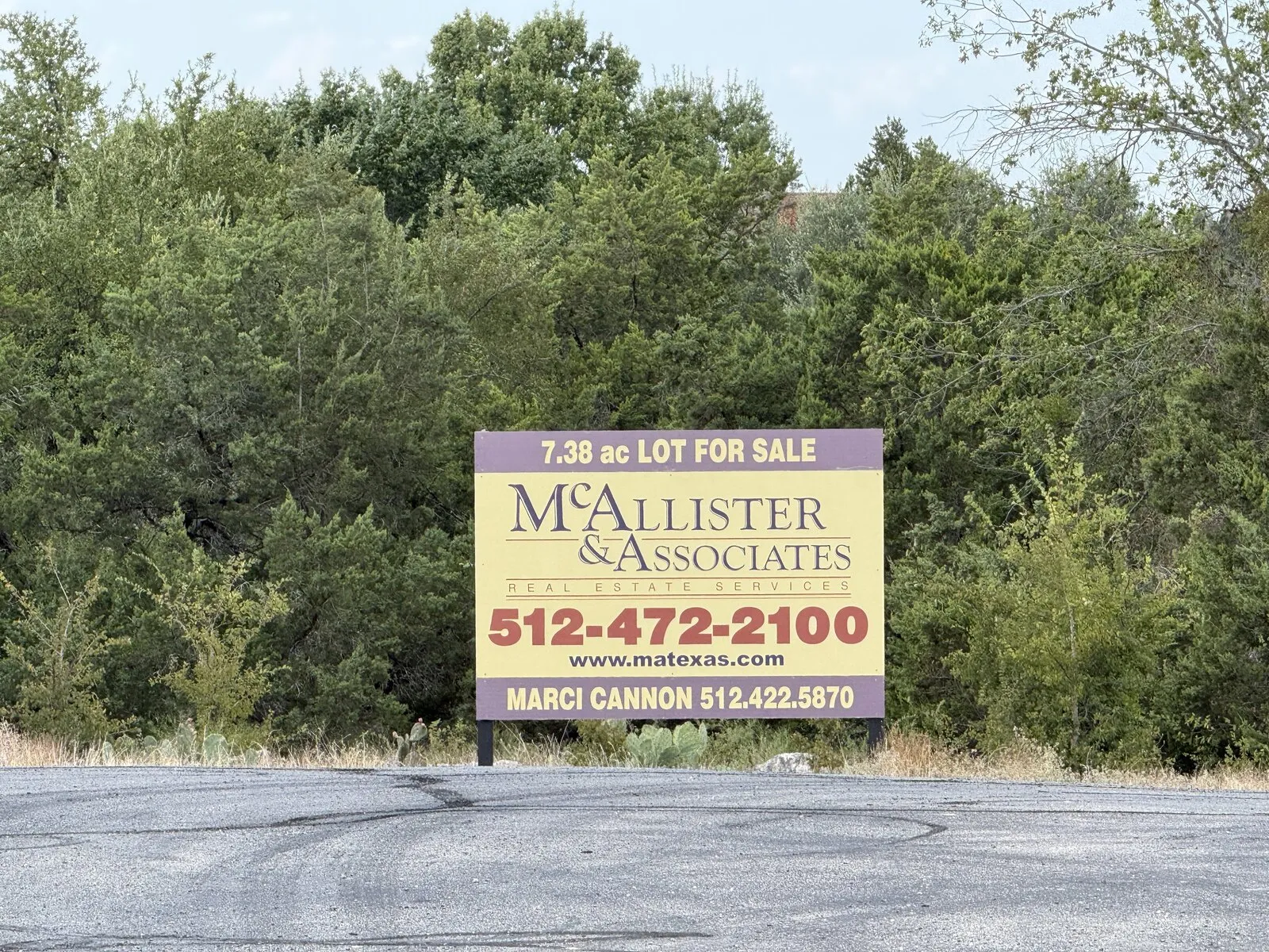 A large yellow and purple real-estate sign reading “7.38 ac LOT FOR SALE — McAllister &amp; Associates Real Estate Services,” with the broker’s phone numbers, standing at the end of the Chitina Court cul-de-sac in front of the park’s cedar and oak trees.