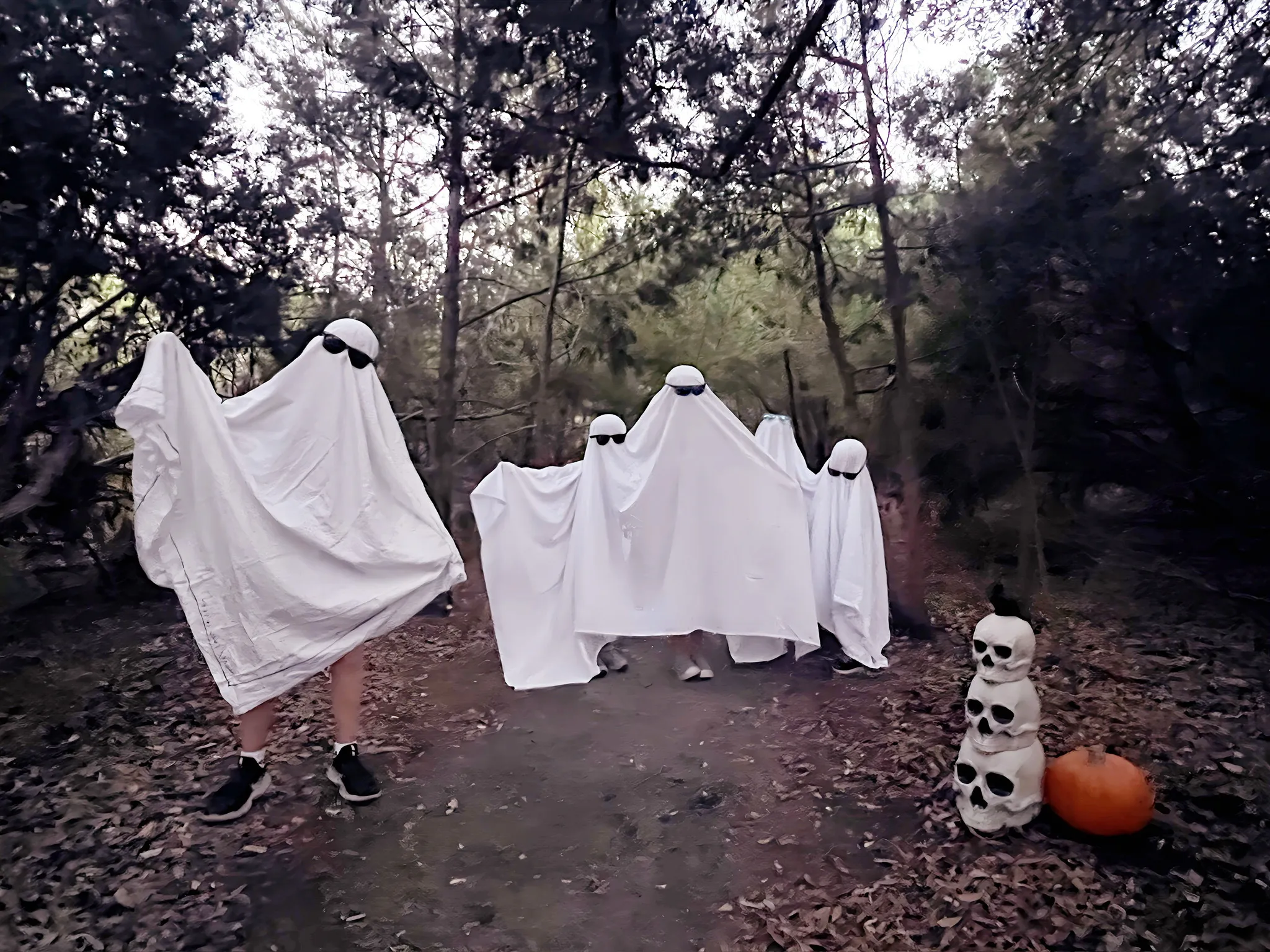 Kids in white-sheet ghost costumes on the trail at Halloween, with a stack of skulls and a pumpkin.